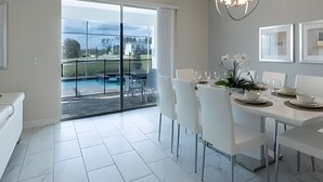 A modern dining area with a glass door leading to a pool, white furniture, and a chandelier. - Spacious 8BR Retreat with Private Pool at CG (Kissimmee)