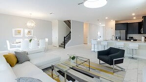 A modern living room with a sofa, coffee table, and a black chair. There is a staircase leading to another level, and a kitchen area with a bar counter. - Spacious 8BR Retreat with Private Pool at CG (Kissimmee)