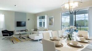 A modern living room with a dining area, a flat-screen TV, and a large window offering a view of the outdoors. - Spacious 8BR Retreat with Private Pool at CG (Kissimmee)