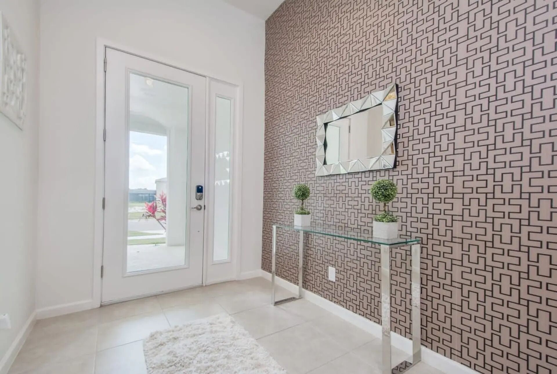 A hallway with a patterned wall, a glass console table with potted plants, and a white door with a glass panel.