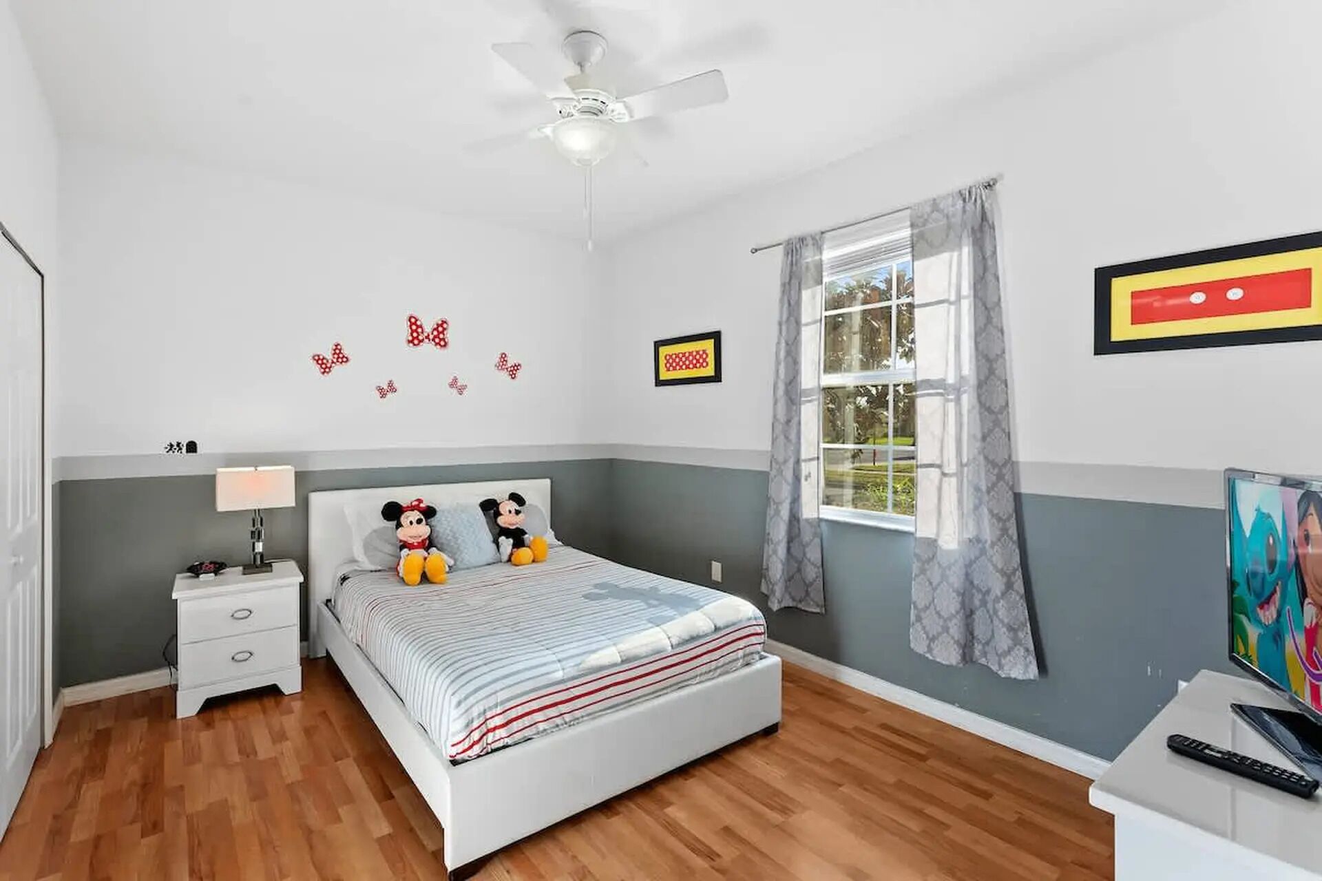 Een slaapkamer met een bed, twee Mickey Mouse knuffels, een nachtkastje, een lamp, een plafondventilator, een raam met gordijnen en een schilderij aan de muur.