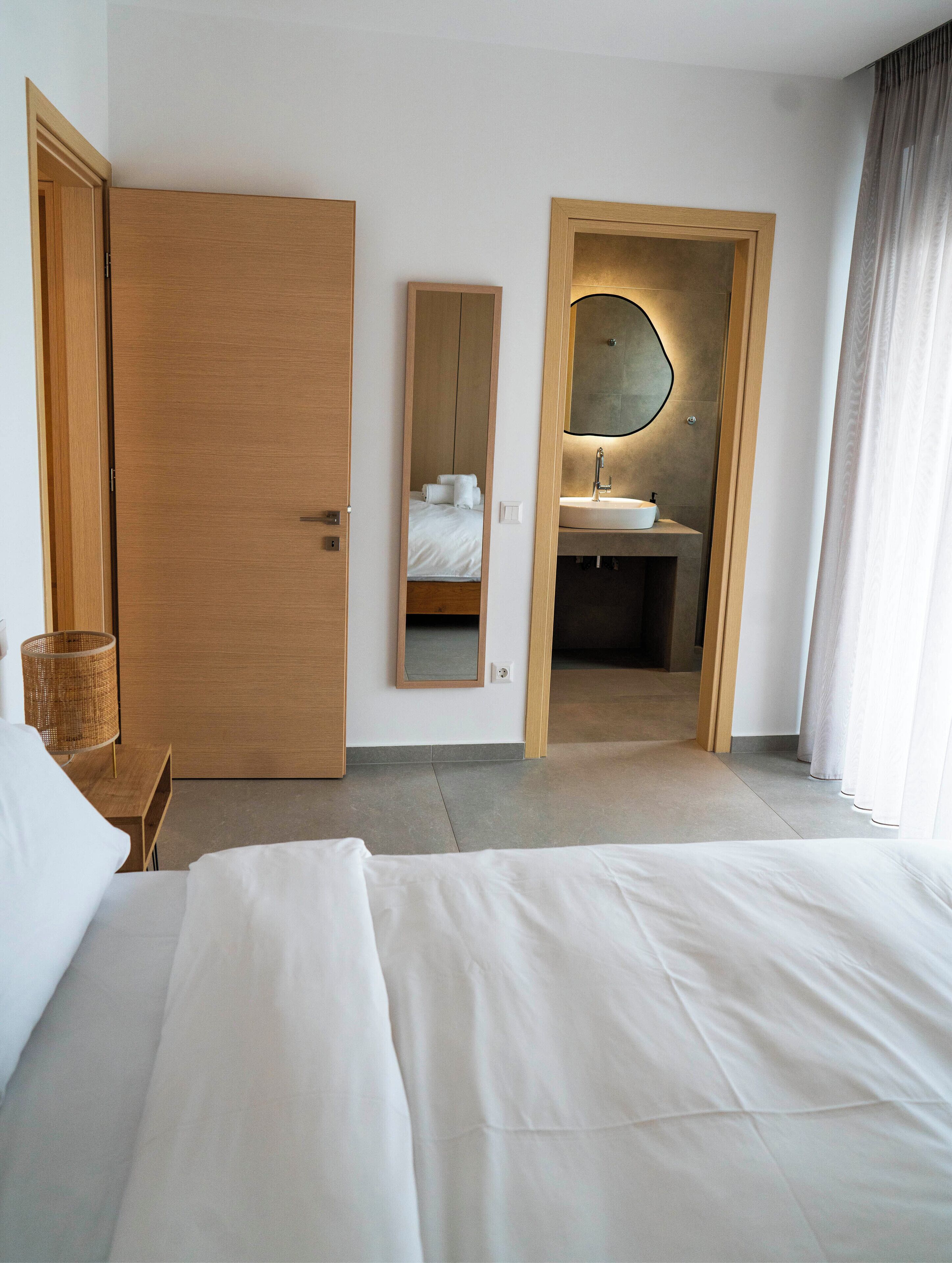 Ein Hotelzimmer mit einem großen Bett, einer Holztür, einem Spiegel und einem durch eine geöffnete Tür einsehbarer Badezimmer.