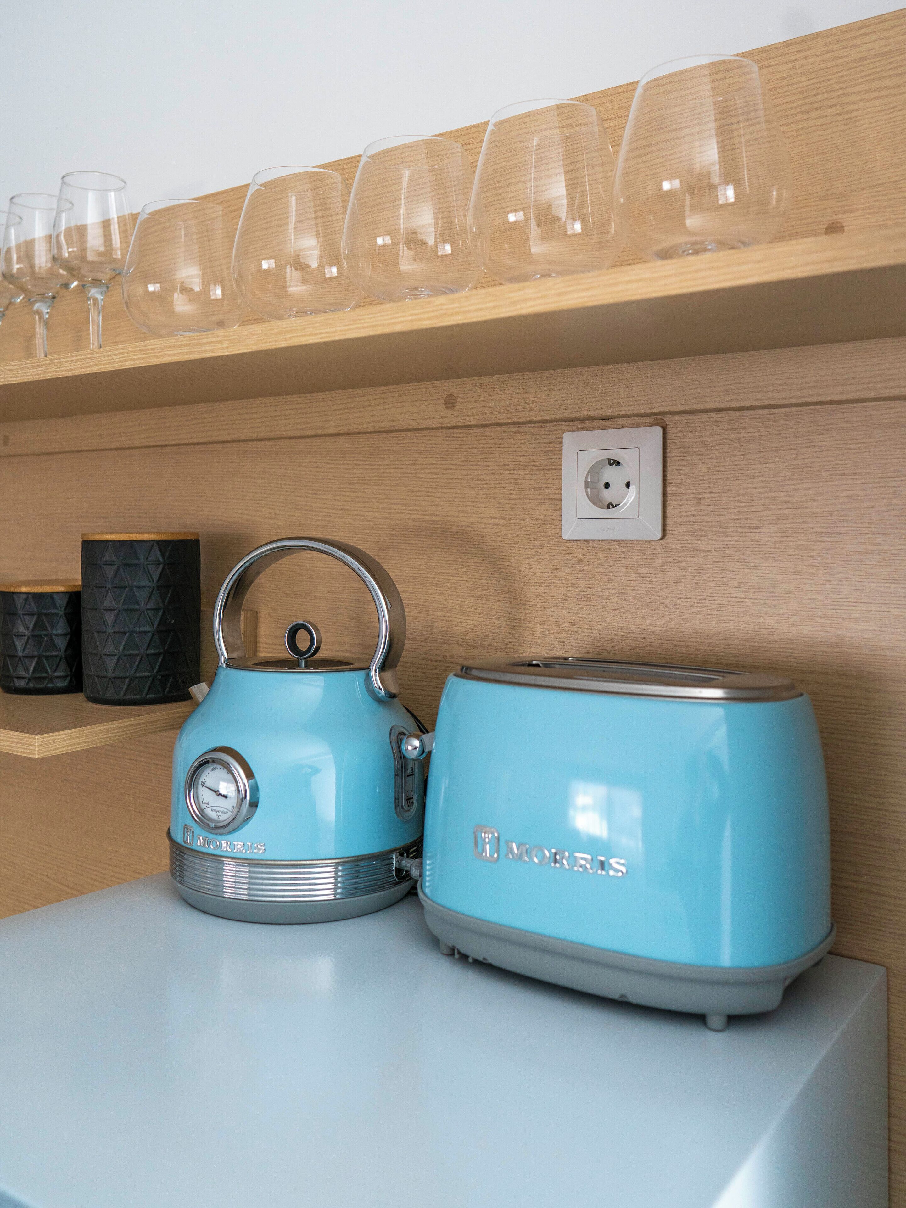 Une cuisine avec une bouilloire et un grille-pain bleus posés sur un plan de travail blanc, une étagère en bois au-dessus où sont rangés des verres à vin, et une prise électrique sur le mur.