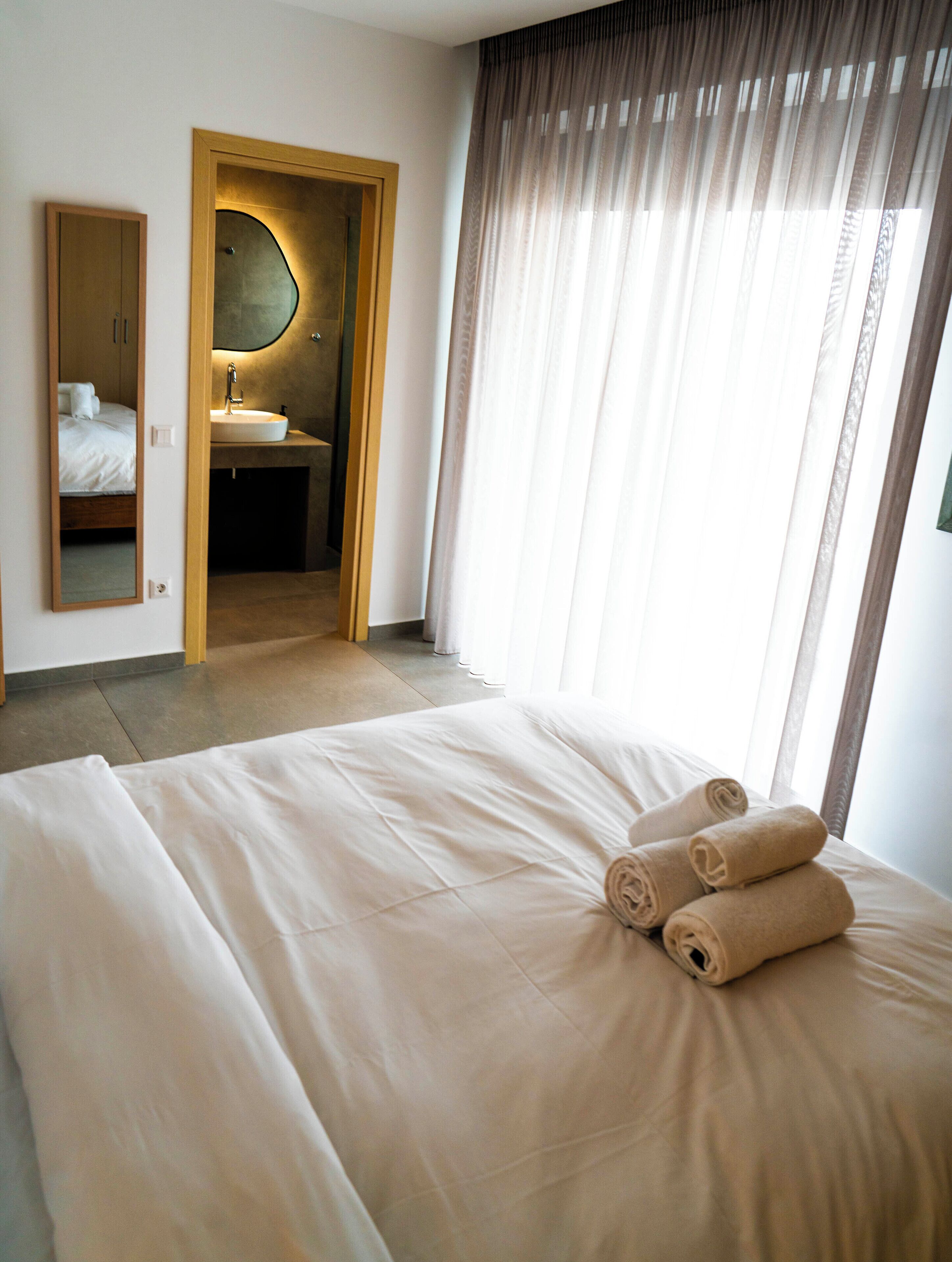 Une chambre d’hôtel avec un grand lit, une salle de bain avec un lavabo et un miroir, et une vue à travers des rideaux transparents.