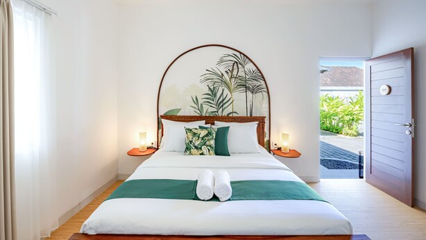 A neatly made bed with two rolled towels, a green throw, and a tropical leaf wall art. There are two bedside lamps, a wooden headboard, and a door leading outside. - Cove Dahayu Sanctuary (Denpasar)