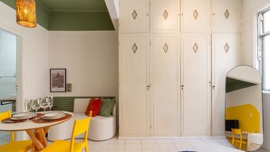 A dining area with a round table, yellow chairs, and a sofa with cushions. There are white cabinets with diamond-shaped handles and a large mirror reflecting part of the room. - Cozy 6 minutes from Copacabana Beach | BR710 (Rio de Janeiro)