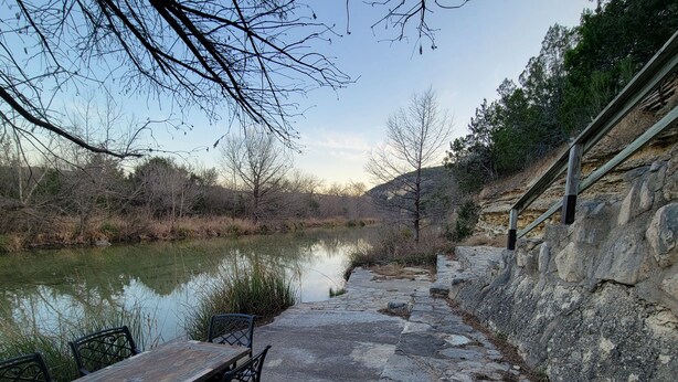 Un sendero de piedra que conduce a una mesa y sillas de madera junto a un río tranquilo, con un muro de piedra en un lado y árboles al fondo.