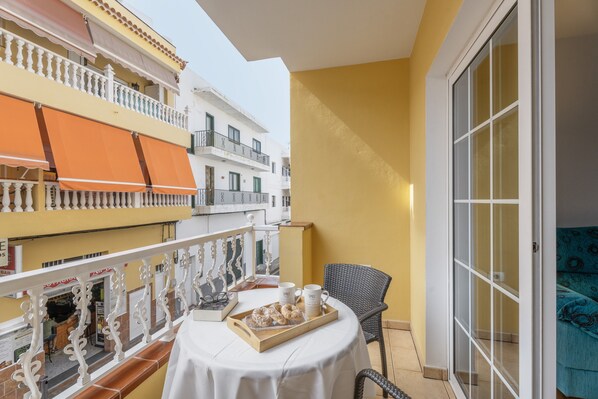 Un balcón con una mesa lista para el desayuno, una silla de mimbre y vistas a edificios cercanos.
