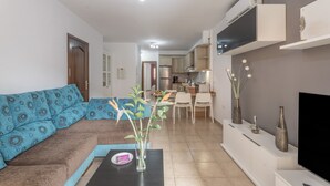 A living room with a sofa, a coffee table with a vase, a TV, and a kitchen in the background.