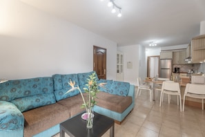 Una sala de estar con un sofá, una mesita con un florero, y una cocina con nevera y armarios.
