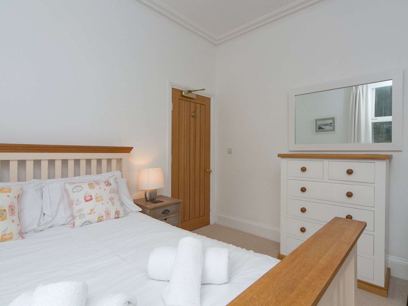 A bedroom with a bed, bedside table, lamp, wooden dresser, and a mirror.