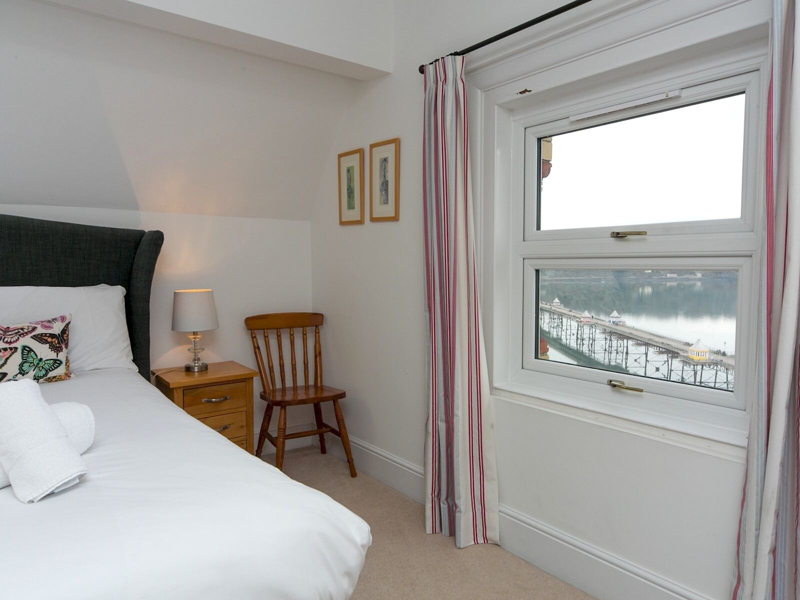 Une chambre à coucher bien rangée, avec un lit, une table de chevet, une lampe, une chaise et une fenêtre donnant sur une jetée et l’eau.