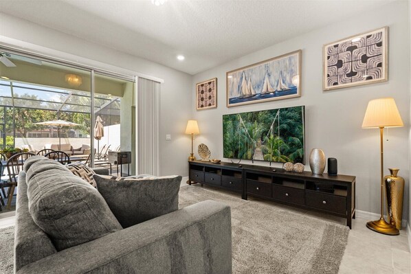 A modern living room with a grey sofa, a TV mounted on the wall, and a dining area with outdoor seating visible through large windows.