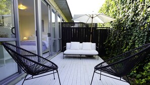 A deck with a white sofa and two black chairs, a large umbrella, and a glass door leading to a bedroom.