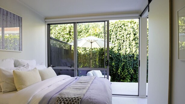 A bedroom with a large bed, a sliding glass door to a balcony, and a view of greenery outside.