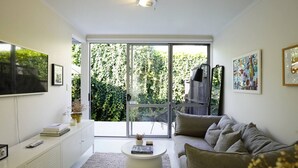 A modern living room with a sofa, a coffee table, and a TV on the wall. There is a sliding glass door leading to a balcony with greenery outside.