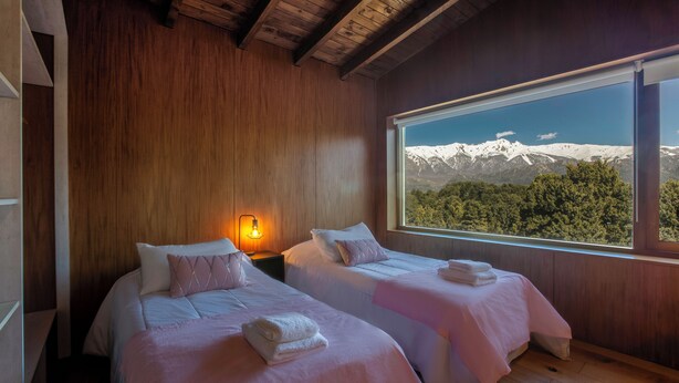A bedroom with two beds, wooden walls, a nightstand with a lamp, and a large window with a mountain view. - Apartments Bariloche - Lago Gutierrez (San Carlos de Bariloche)
