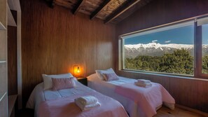 Un dormitorio con dos camas, paredes de madera, una mesita de noche con lámpara y un ventanal con vistas a la montaña.