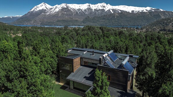 A modern house with a sloped roof, solar panels, and a view of snow-capped mountains and a lake.