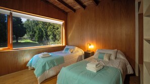 A room with two beds, wooden walls, a window with a view of trees, and a nightstand with a lamp.
