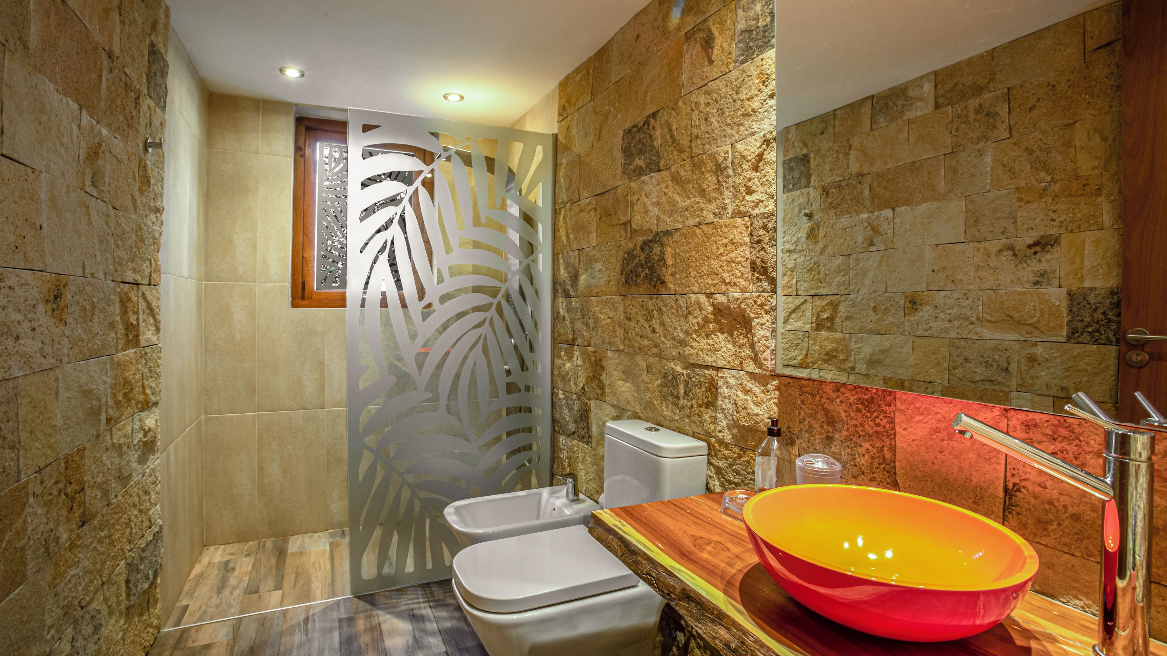 Un bagno con parete in pietra, mobile bagno in legno, lavabo a forma di coppa rosso e un water.