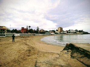Una playa con una persona caminando, una cerca blanca y edificios al fondo.