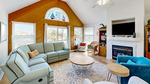 Een ruime woonkamer met een hoge plafond, een open haard, een televisie erboven en een gezellige zithoek met een bank en fauteuils.