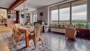 Un comedor con mesa de madera y sillas de mimbre, vistas a través de ventanas amplias y un balcón con plantas en macetas.