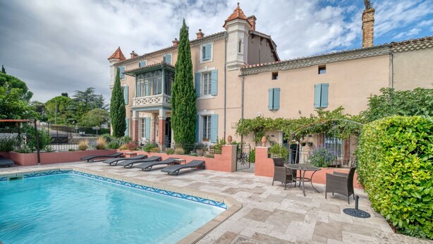 Una piscina con tumbonas, un patio con mesas y sillas, y un edificio con una torre y varias ventanas.