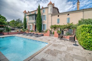 A swimming pool with lounge chairs, a patio area with tables and chairs, and a building with a tower and multiple windows. - Holiday Home 'Château' with Mountain View, Balcony & Wi-Fi (Souilhanels)