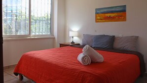 A neatly made bed with a red bedspread and a rolled towel, a nightstand with a lamp, and a painting on the wall. - Stay 5 min from the beach and downtown Cancun. (Cancún)