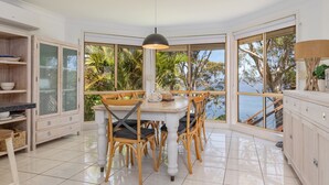 A dining area with a table and chairs, large windows with a view of trees and water, and light-colored cabinetry. - Luminara Luxury Lakefront (Smiths Lake)