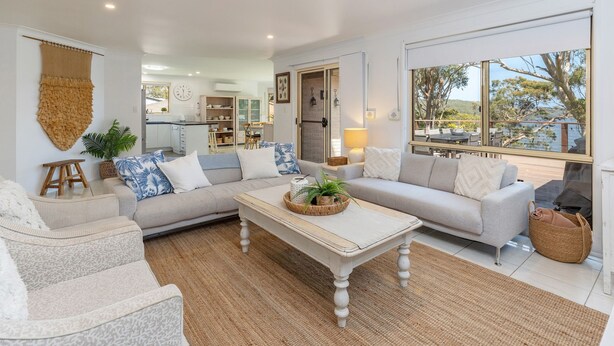 A spacious living room with a large sectional sofa, a coffee table, and a view of the outdoors through large windows. - Luminara Luxury Lakefront (Smiths Lake)