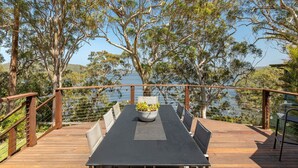 A wooden deck with a table and chairs, overlooking a body of water and surrounded by trees. - Luminara Luxury Lakefront (Smiths Lake)