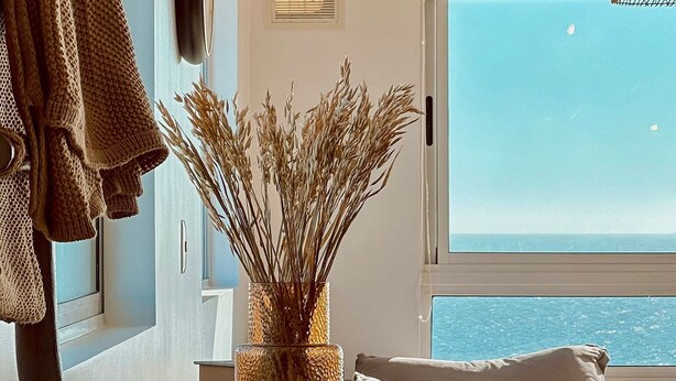 Ein Wohnzimmer mit Meerblick, ein weißes Sofa, ein Holztisch mit einer Vase getrockneter Pflanzen und eine hängende Leuchte.