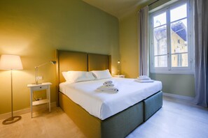 Una cama bien hecha con sábanas y toallas blancas, una lámpara de noche y una ventana con cortinas.