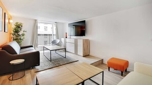 A modern living room with a sofa, coffee table, and ottoman. A flat-screen TV is mounted on the wall, and there is a window with a view of buildings. - Charming Apartment- 3br-8p Opera (Paris)