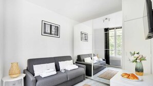 A modern hotel room with a grey sofa, a side table with a lamp, a large mirror, and a vase with flowers. - Studio in the Heart of the Marais (Paris)