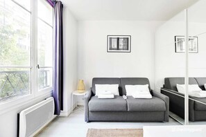 A modern, minimalist hotel room with a grey sofa, a small side table with a lamp, and a large window with a view of buildings and greenery. - Studio Cosy- 2P- of the Marais (Paris)