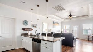 A spacious kitchen with white cabinetry, a granite countertop, and a dishwasher. There is a dining area with a dark sofa and a ceiling fan.