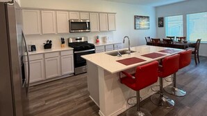Une cuisine moderne avec des meubles blancs, un îlot central doté de tabourets de bar rouges, des appareils en acier inoxydable et un coin repas avec une table et des chaises.