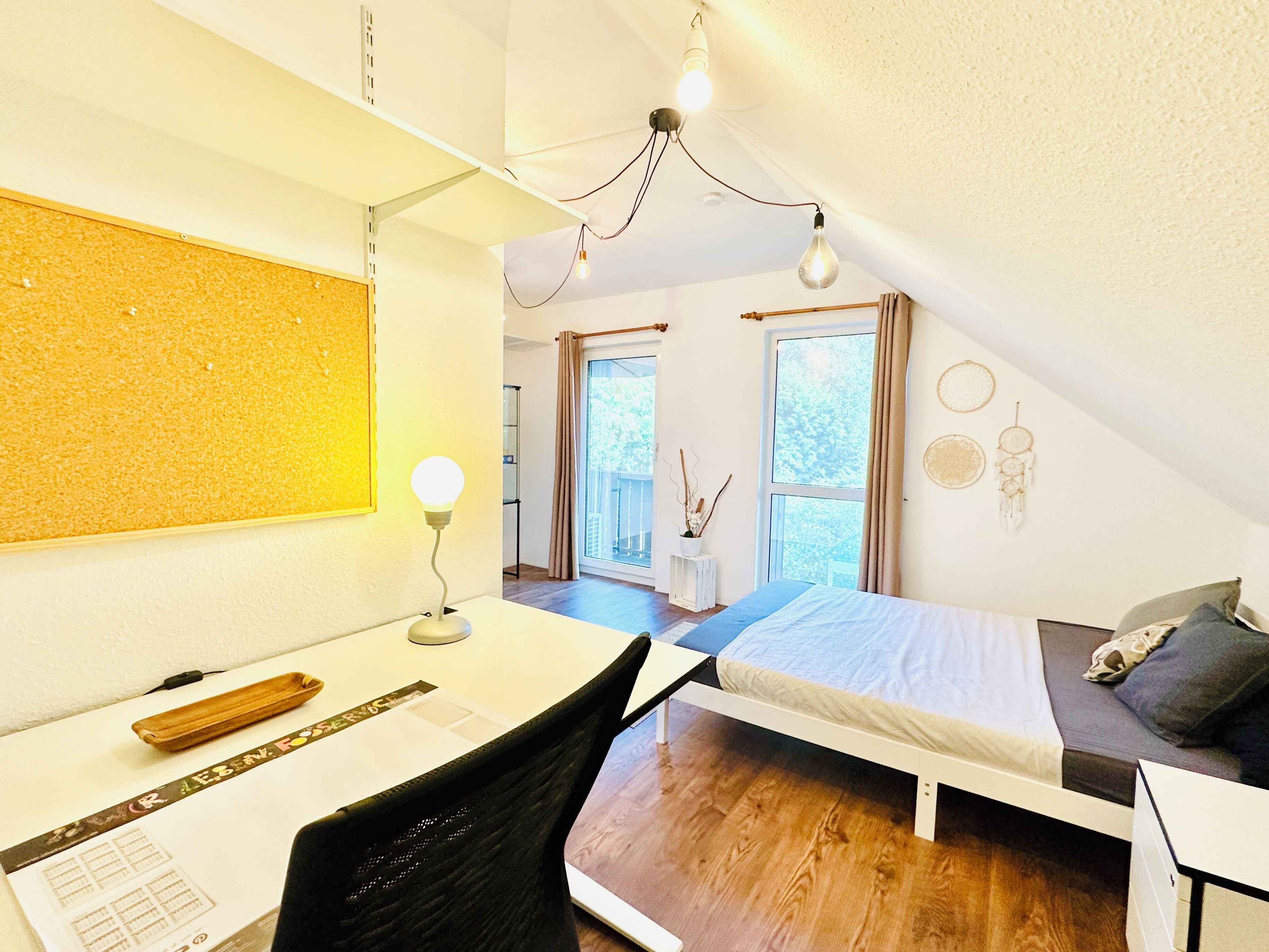 Ein Schlafzimmer mit Bett, Schreibtisch, Stuhl und Blick ins Freie durch ein Fenster.