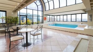 An indoor swimming pool with a view of the city, surrounded by tiled flooring and metal chairs. - Meu Anfitrião Nações Unidas (São Paulo)