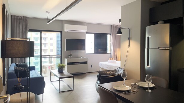 A modern living room with a sofa, a dining table set for two, a TV mounted on the wall, and a large window with curtains. - Meu Anfitrião Nações Unidas (São Paulo)