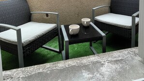 A small outdoor seating area with two wicker chairs and a table, a small window, and a building with a number '21' sign in the background.