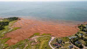 An aerial view of a coastal area with a large, reddish-brown sandbar extending into the water, surrounded by greenery and residential buildings.