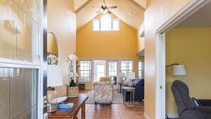 A spacious living room with a high ceiling, a ceiling fan, and large windows allowing natural light. There is a comfortable seating area with a sofa and a chair.
