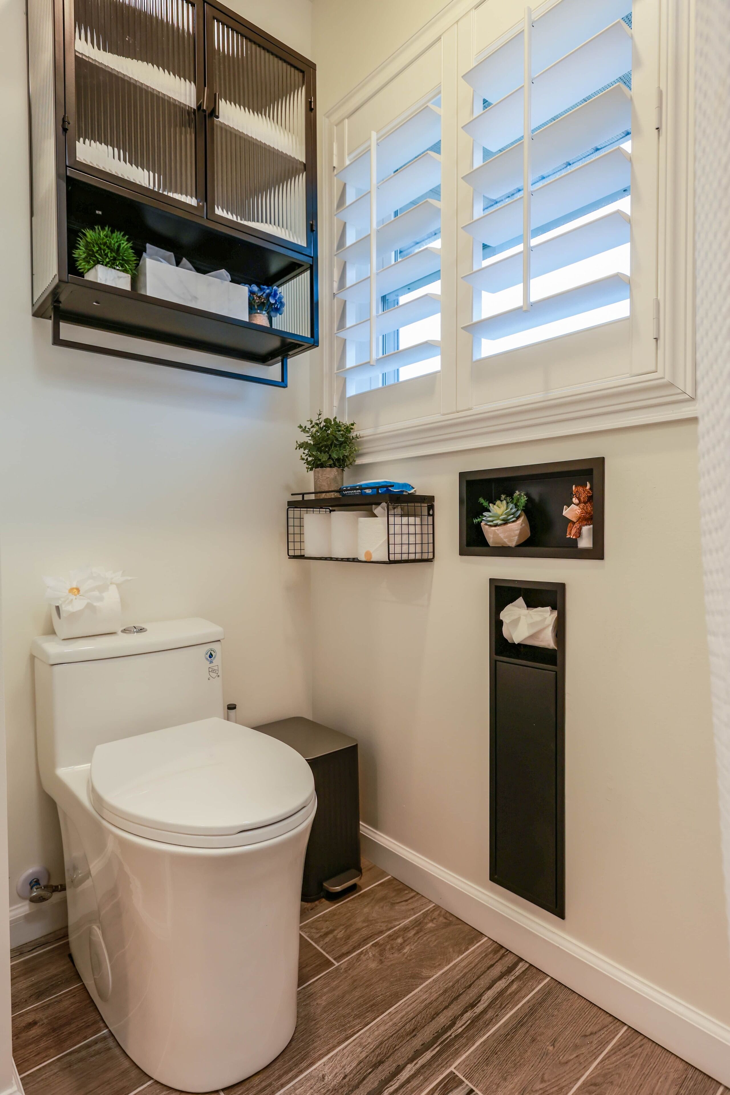 A bathroom with a toilet, a trash bin, a shelf with plants, and a window with blinds.