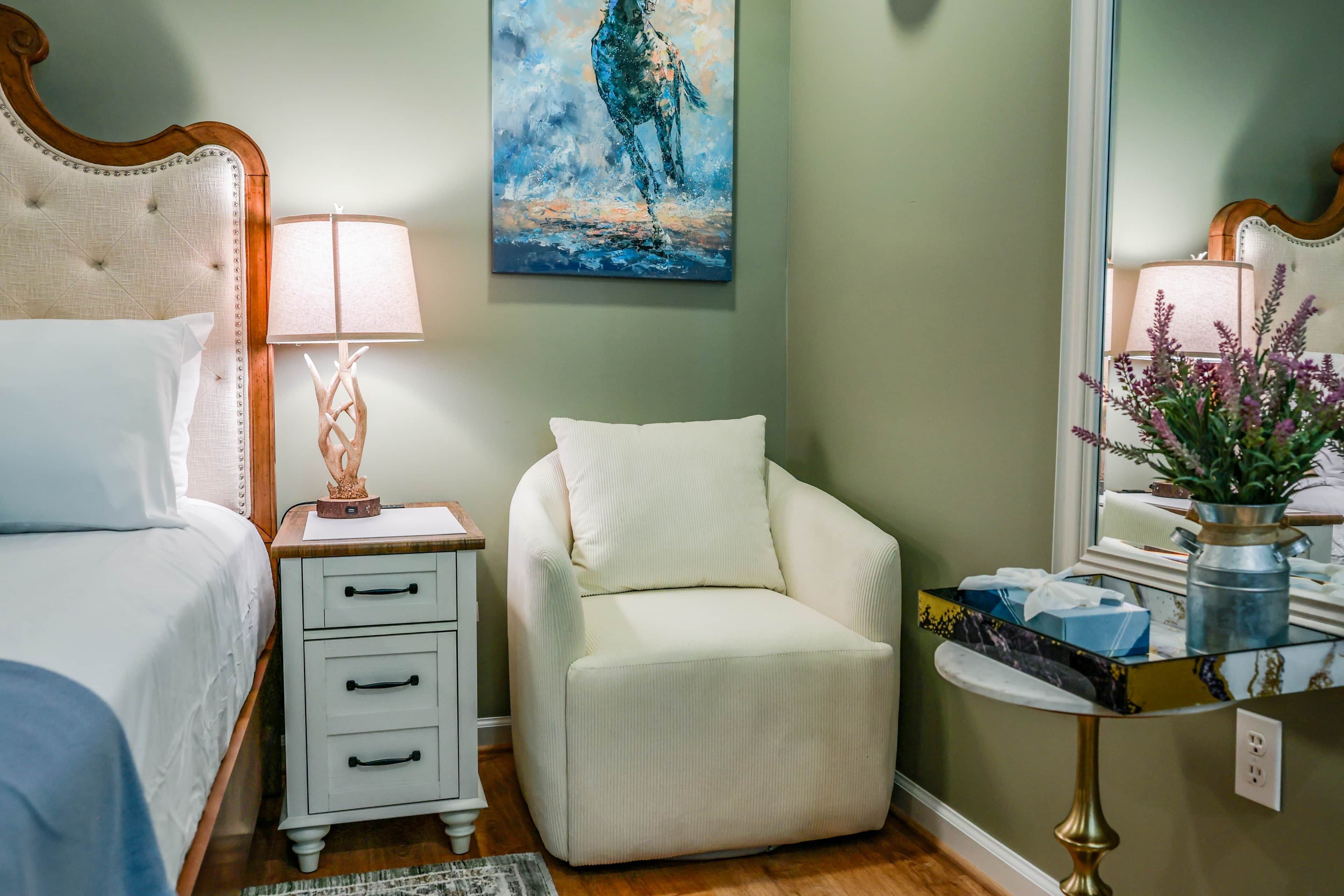 A bedroom with a bed, nightstand, chair, and bedside table with a lamp and decorative items.