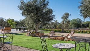 Un patio al aire libre con mesas y sillas, rodeado de un césped cuidado y olivos.